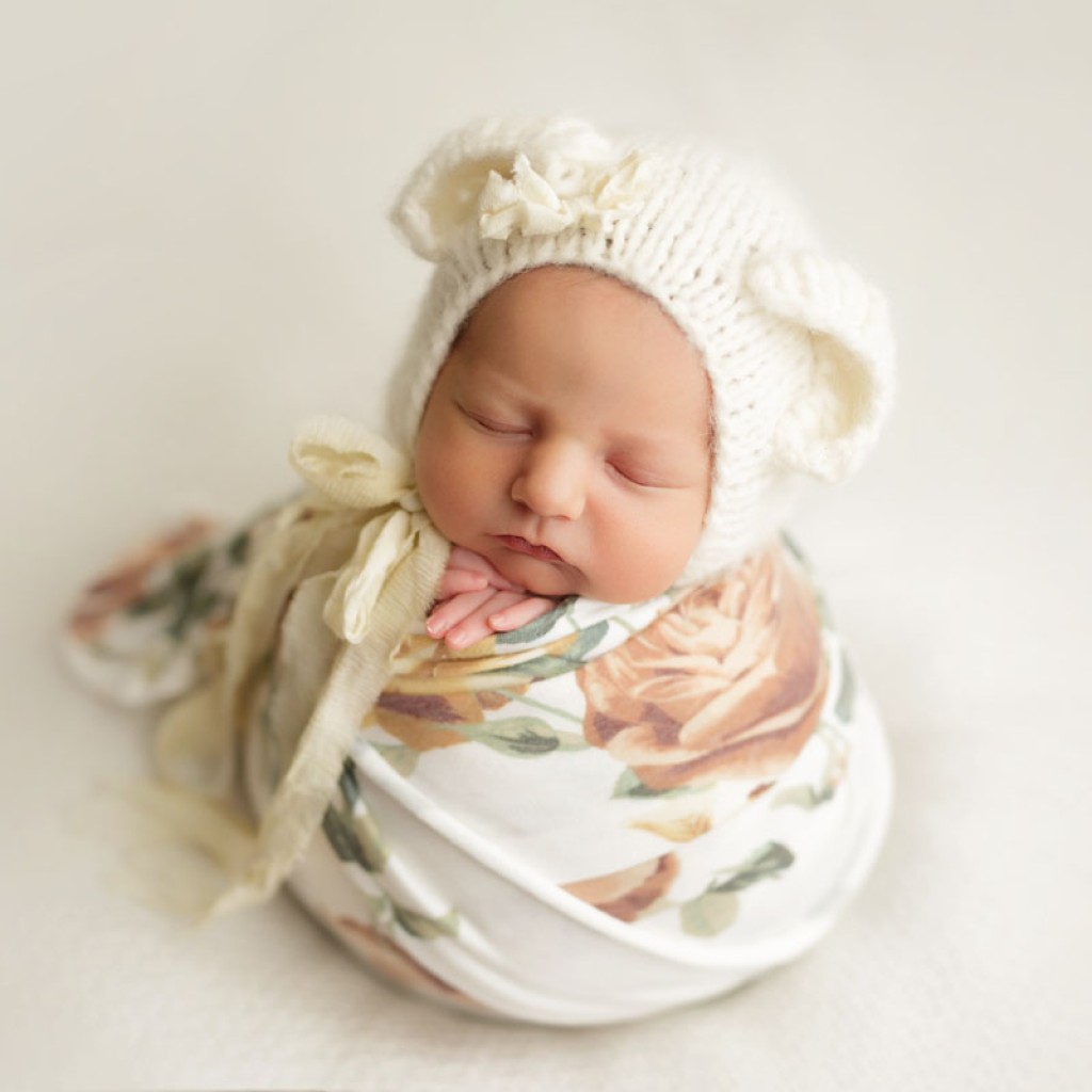 Baby with a floral wrap and teddy bear hat. This is considered a wrapped pose. Sometimes referred to as the potato sack pose or tater tot pose.