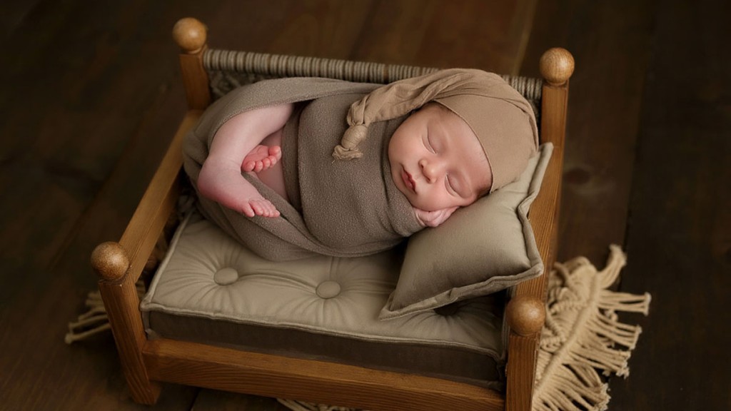 Baby boy wrapped in a brown wrap and wearing a sleepy hat. He is asleep on a pillow on a tiny wooden bed, posed in the San Marcos newborn photography studio by professional photographer Shelly Rosen.