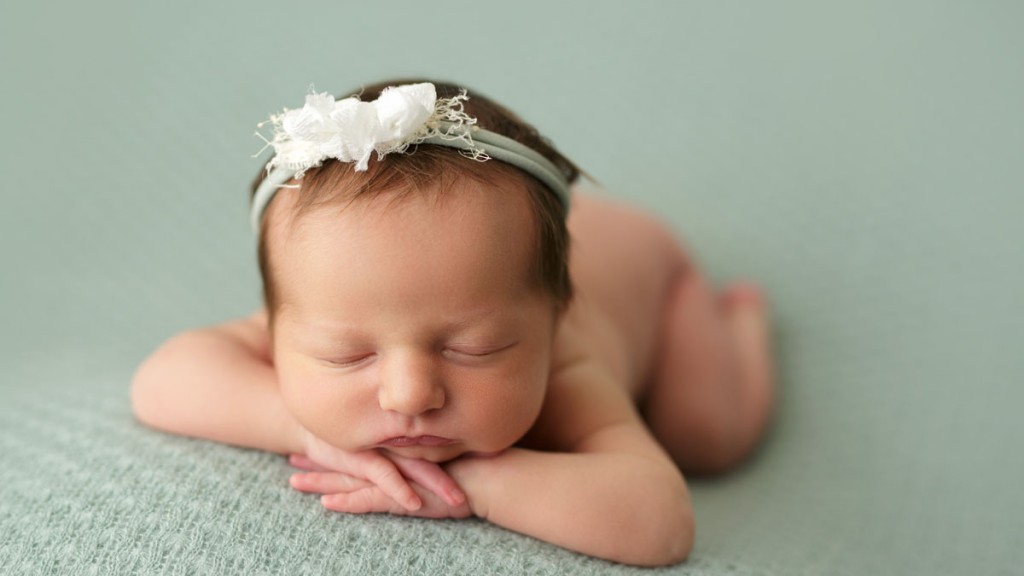 Baby girl with dark hair has a cream bow in her hair and is posed forward facing on a sage green backdrop.